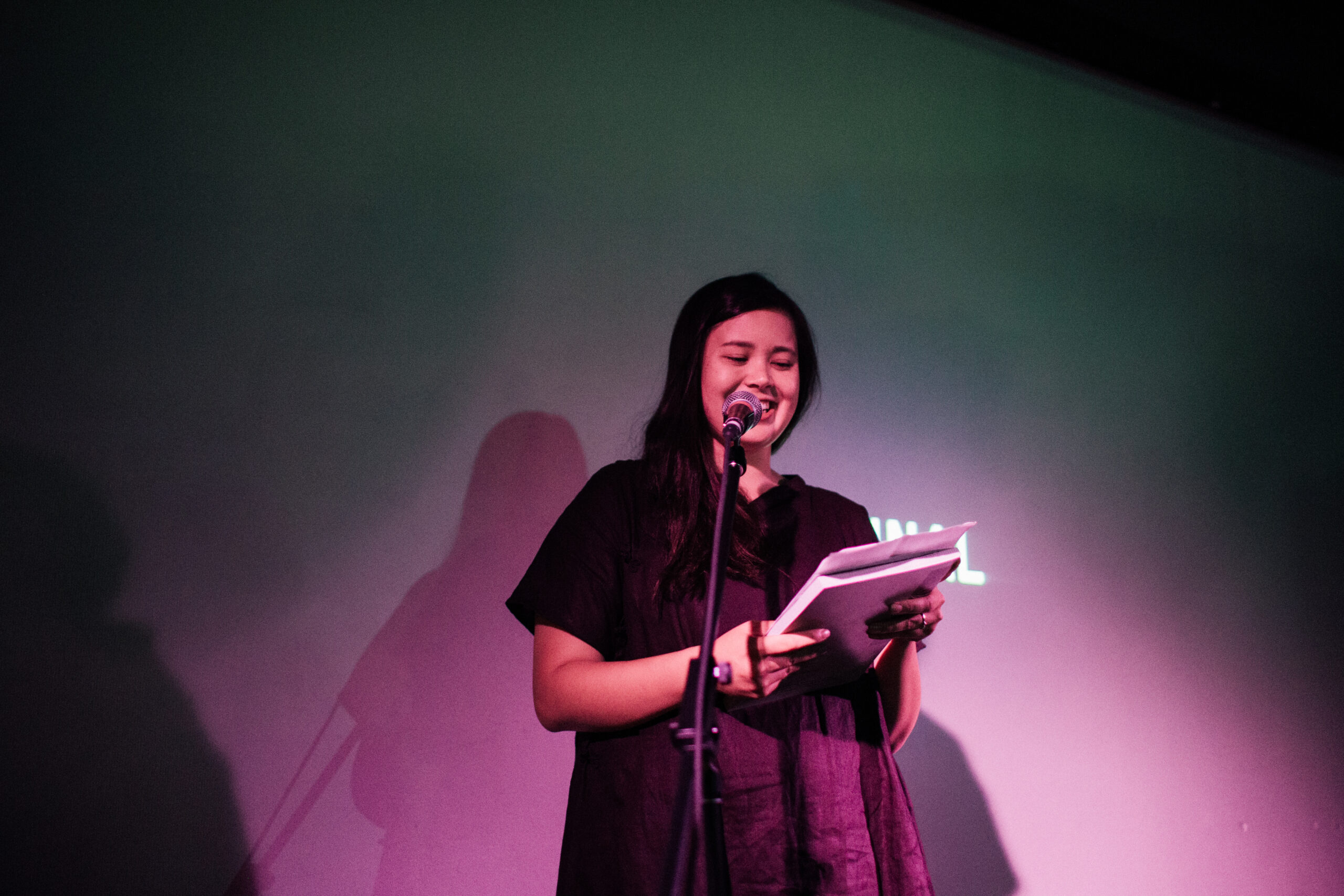 Leah Jing McIntosh looking radiant and smiling as she reads from some papers on top of a book, she is standing on stage in front of a microphone stand in multihued light. Leah has dark, black hair and is wearing flowy dark coloured linen clothing.