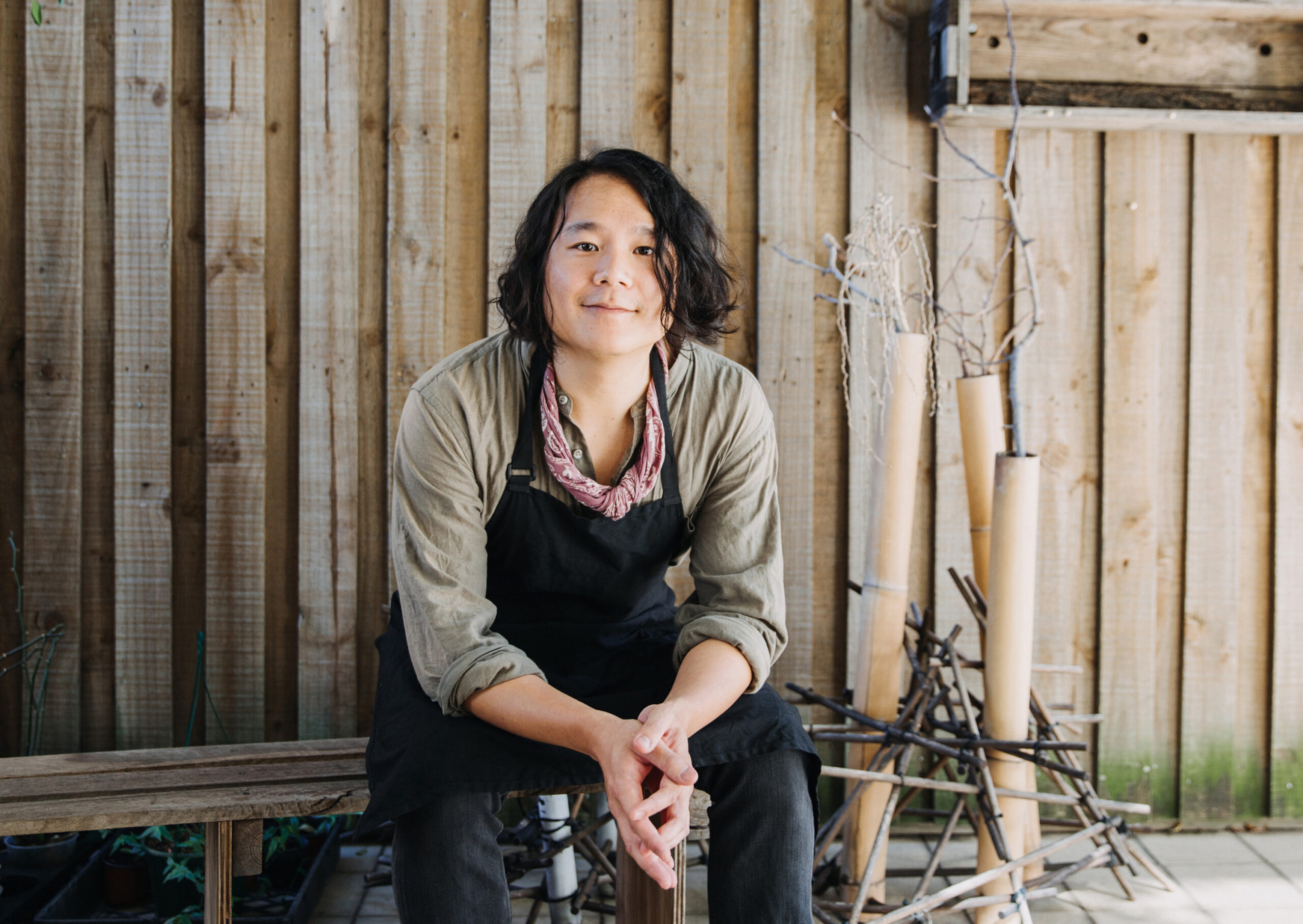 Matt Hsu looking into the camera surrounded by a wood fence and on a wood bench, there are brambles and branches sticking out of tubes and sticks scaffolding them. He is wearing a worker's apron and is sitting, looking warmly into the camera