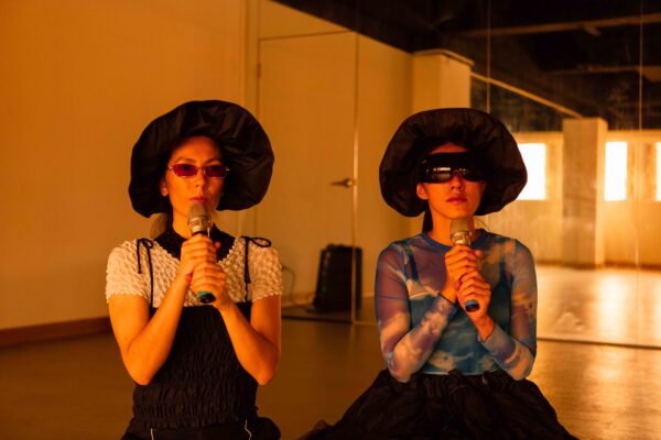 A photo from Paeonia Drive, which features Su Yu Shin and Angela Goh, holding microphones, wearing floppy and wide brimmed hats and funky sunglasses and visors. The room is bathed in orange light and there is a mirror behind them. It looks like they are in a studio or rehearsal or performance space.