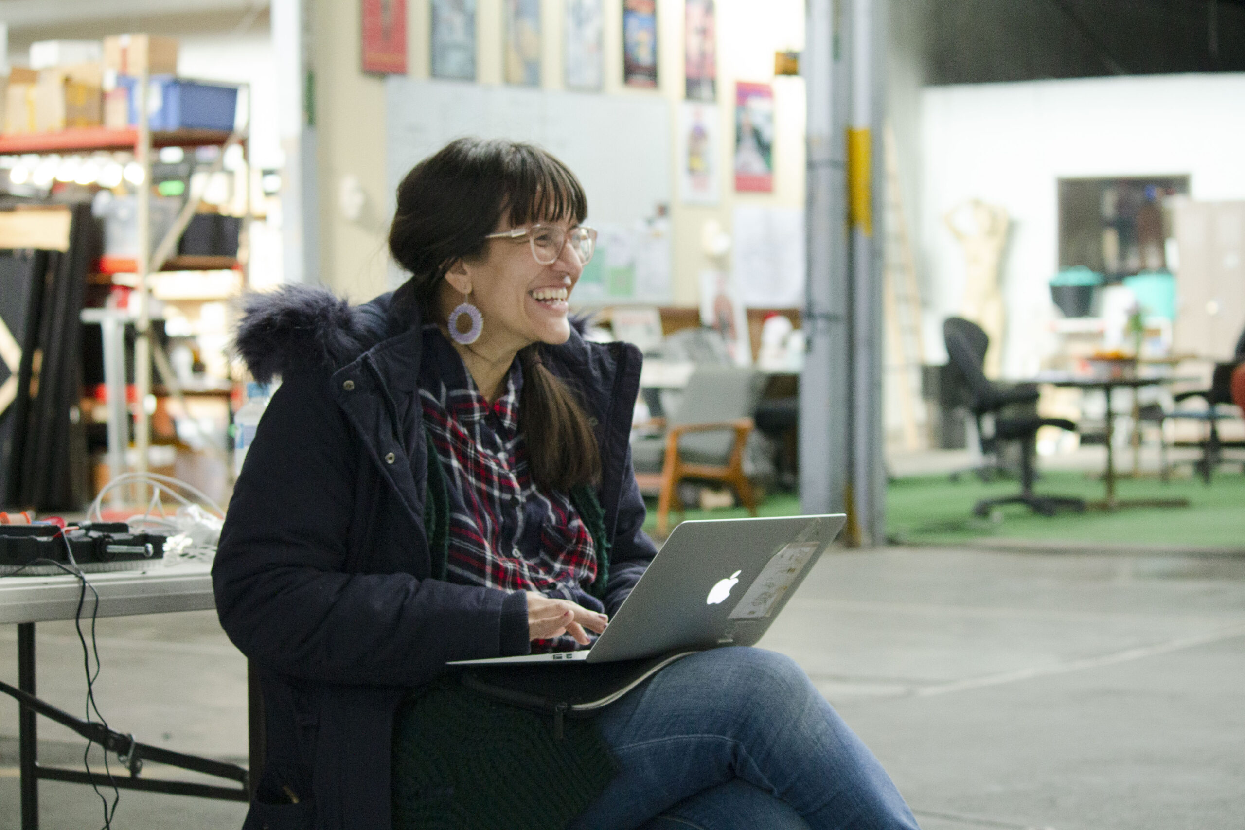 A photo of Yasmin Gurreeboo sitting, with laptop in a backstage theatre lot area, looking happy and productive at work
