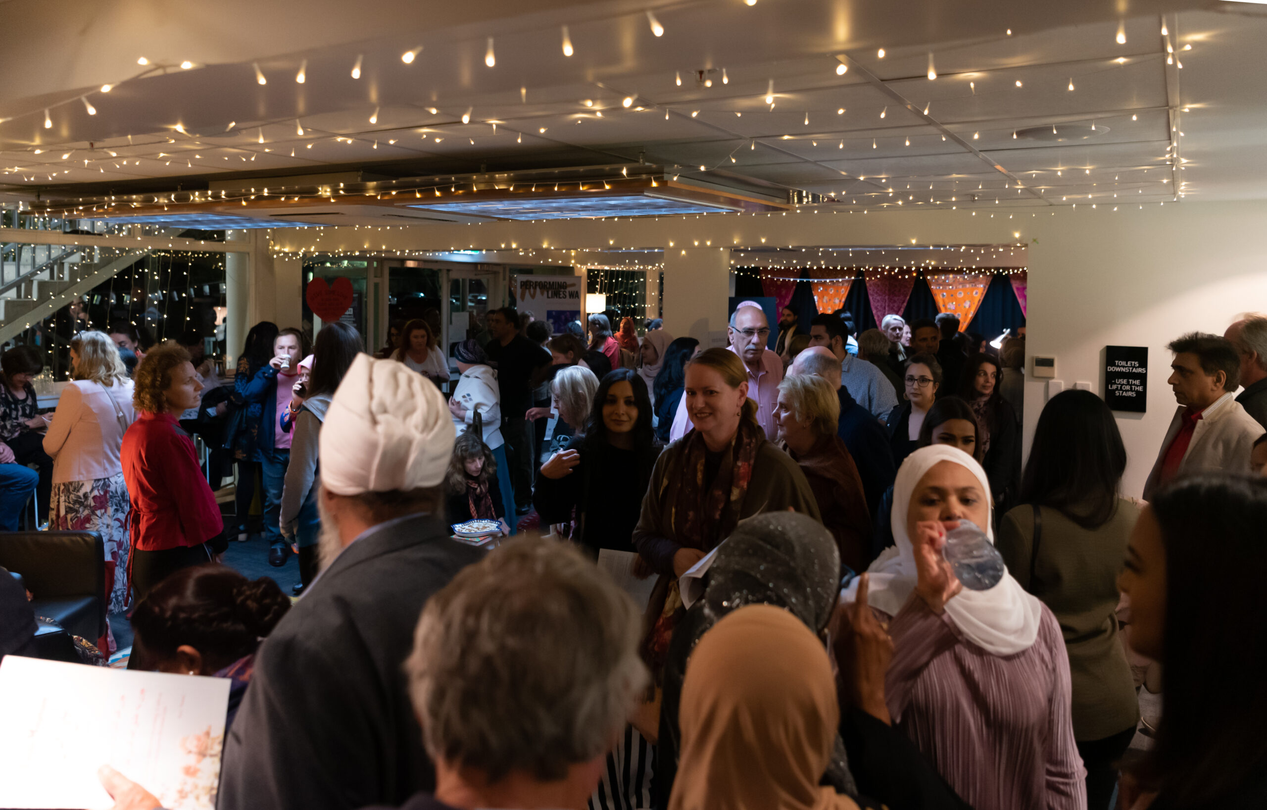A shot of the audience at the Layla Majnun opening night, there is homey lighting around, casting dappled shadows onto the ceiling, the atmosphere is warm and cosy and the crowd is diverse with some members of the crowd wearing turbans and hijabs